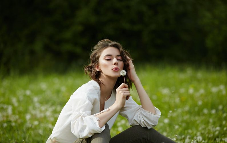 Portrait of a girl sitting in a field on the spring grass among dandelion flowers. Cheerful girl enjoys Sunny spring weather. Natural beauty of a woman, natural cosmetics